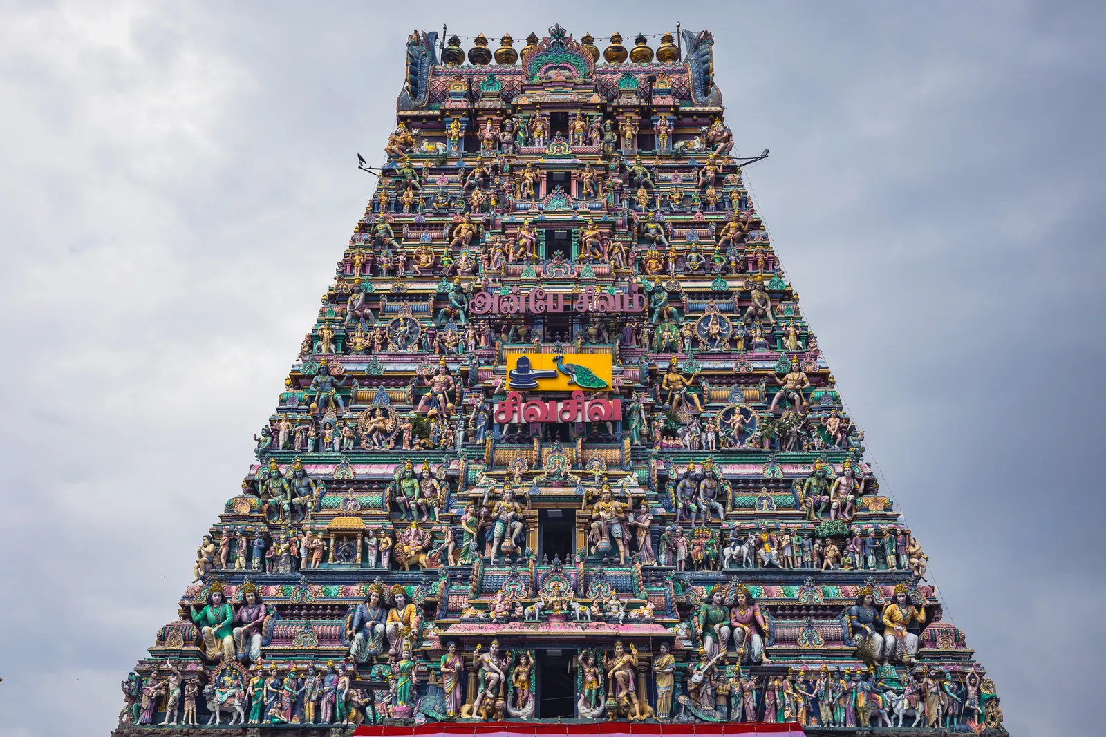 A tall, many-tiered South Indian temple gateway tower covered in colourful sculpted figures, with Tamil lettering on its face, against a grey cloudy sky.