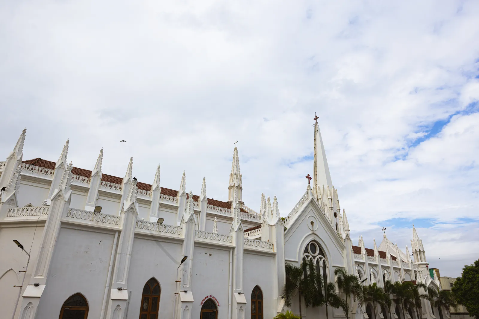 A white Gothic-style church seen from the side, with rows of pointed spires, arched windows and a red-tiled roof, under a cloudy sky with palm trees at the bottom.