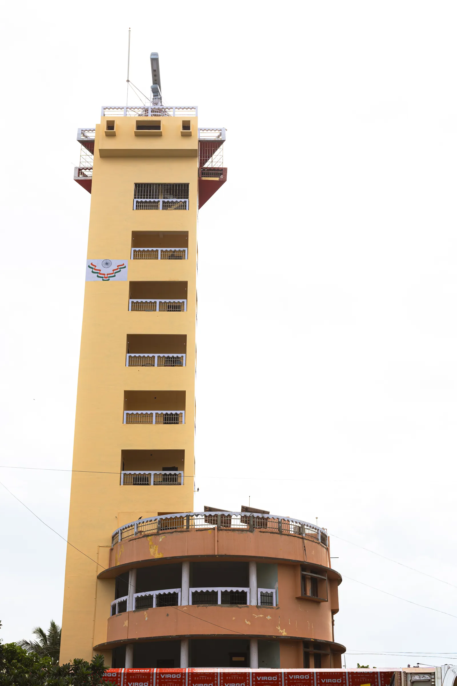 A tall, narrow yellow-ochre tower with a stack of balconies and a lookout platform on top, rising above a curved orange building against a blank white sky.