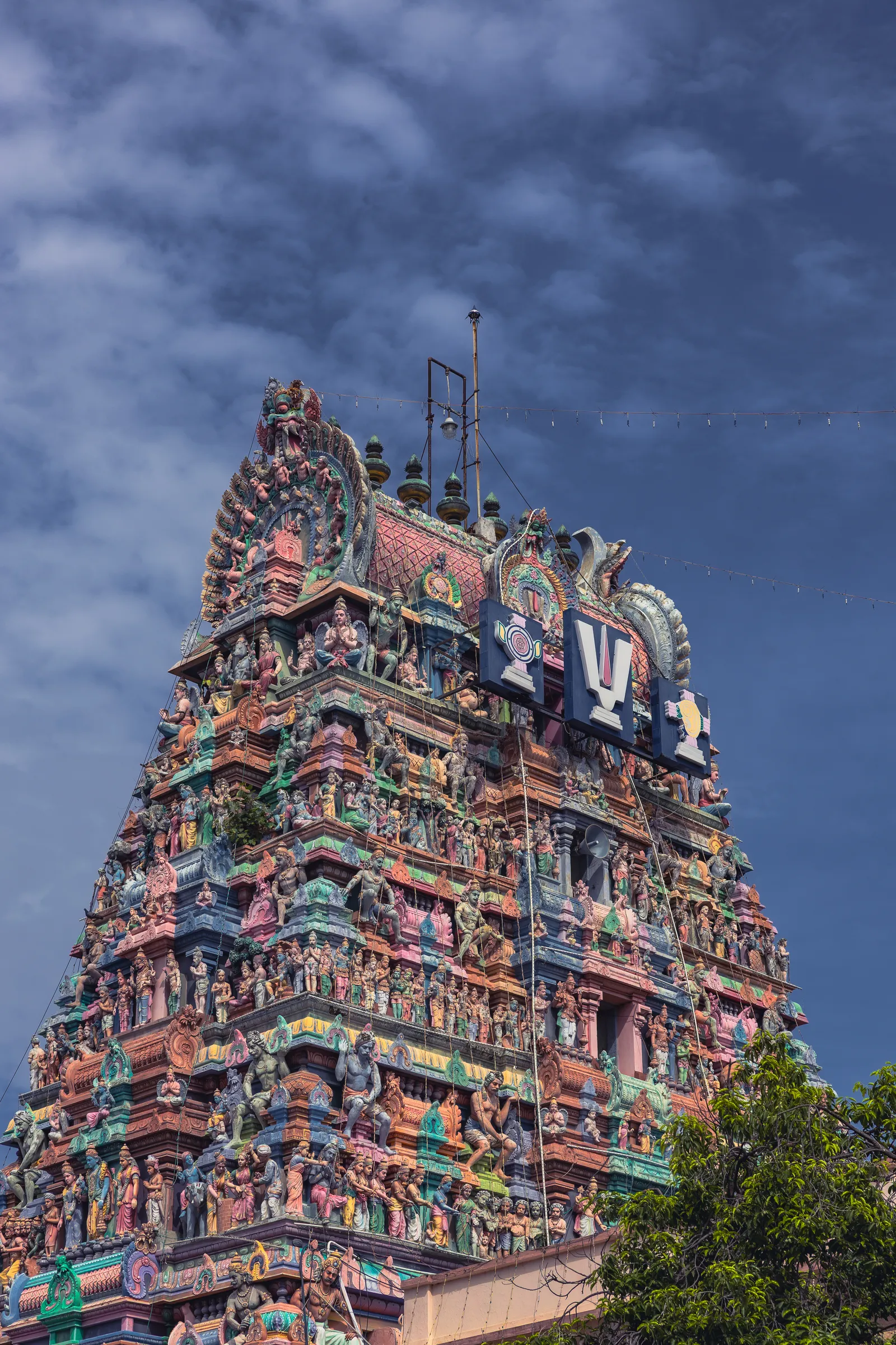The top of a temple tower packed with brightly painted sculptures in pinks, greens and blues, against a dark, stormy blue sky, with a tree in the corner.