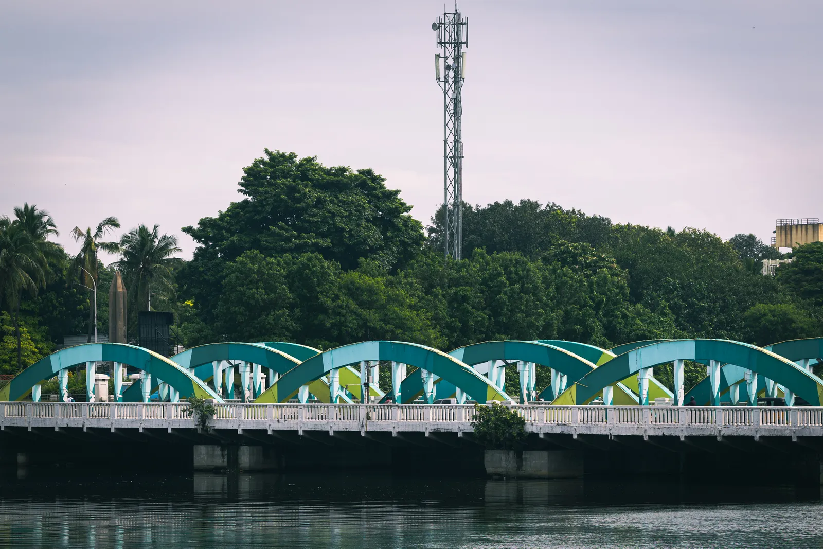 A bridge with a row of teal and green arches over still water, with dense trees and a mobile-phone mast behind it.