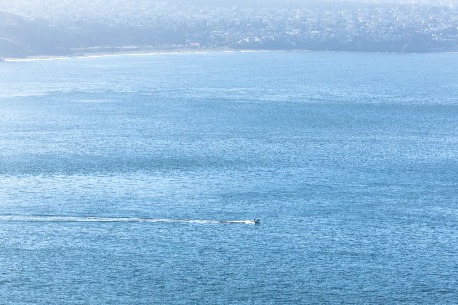 A small motorboat leaving a long white wake across a wide stretch of calm blue water, with a hazy city and coastline on the far shore.
