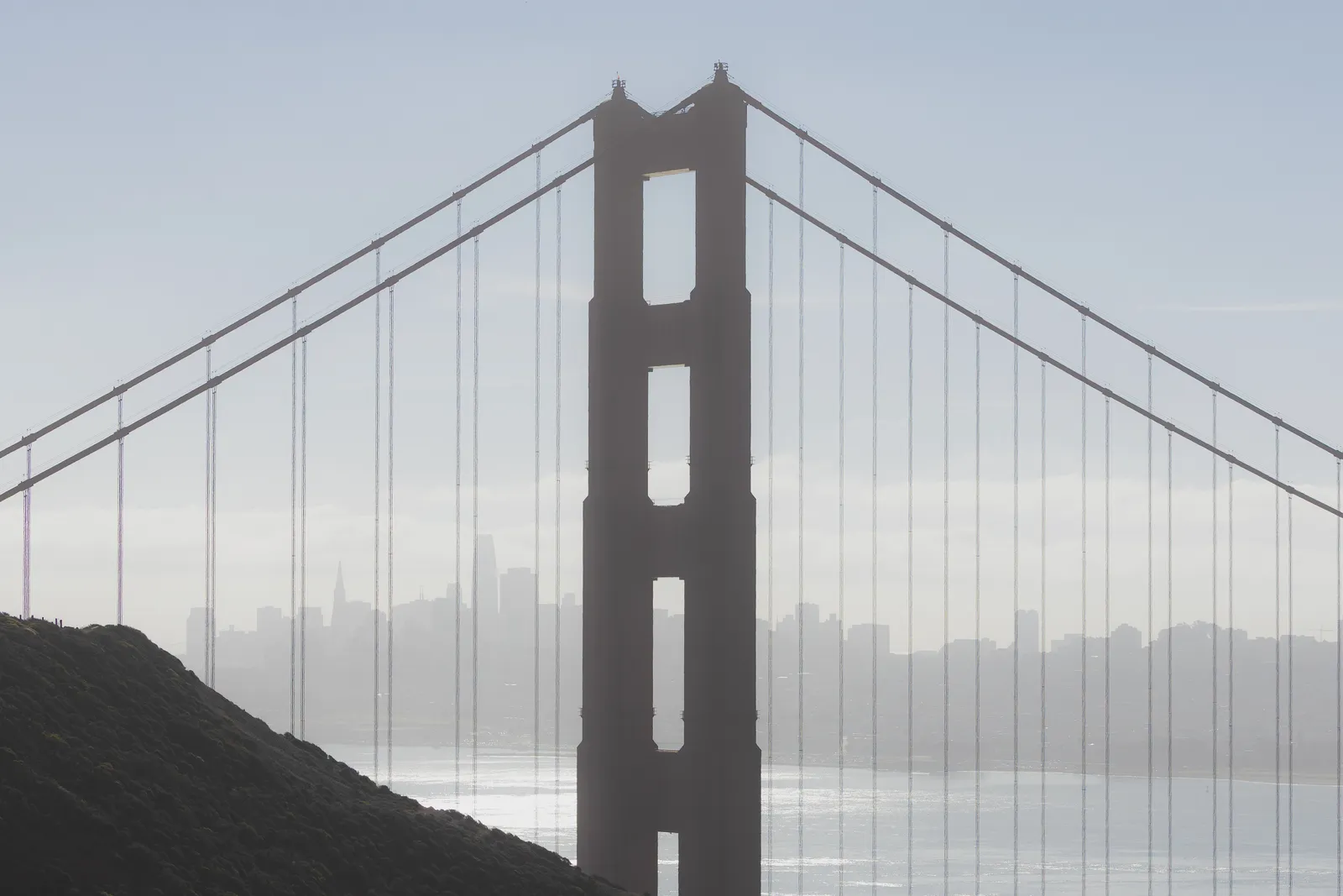One tower of the Golden Gate Bridge with its cables fanning out on both sides, in soft haze, with the San Francisco skyline faint behind it.
