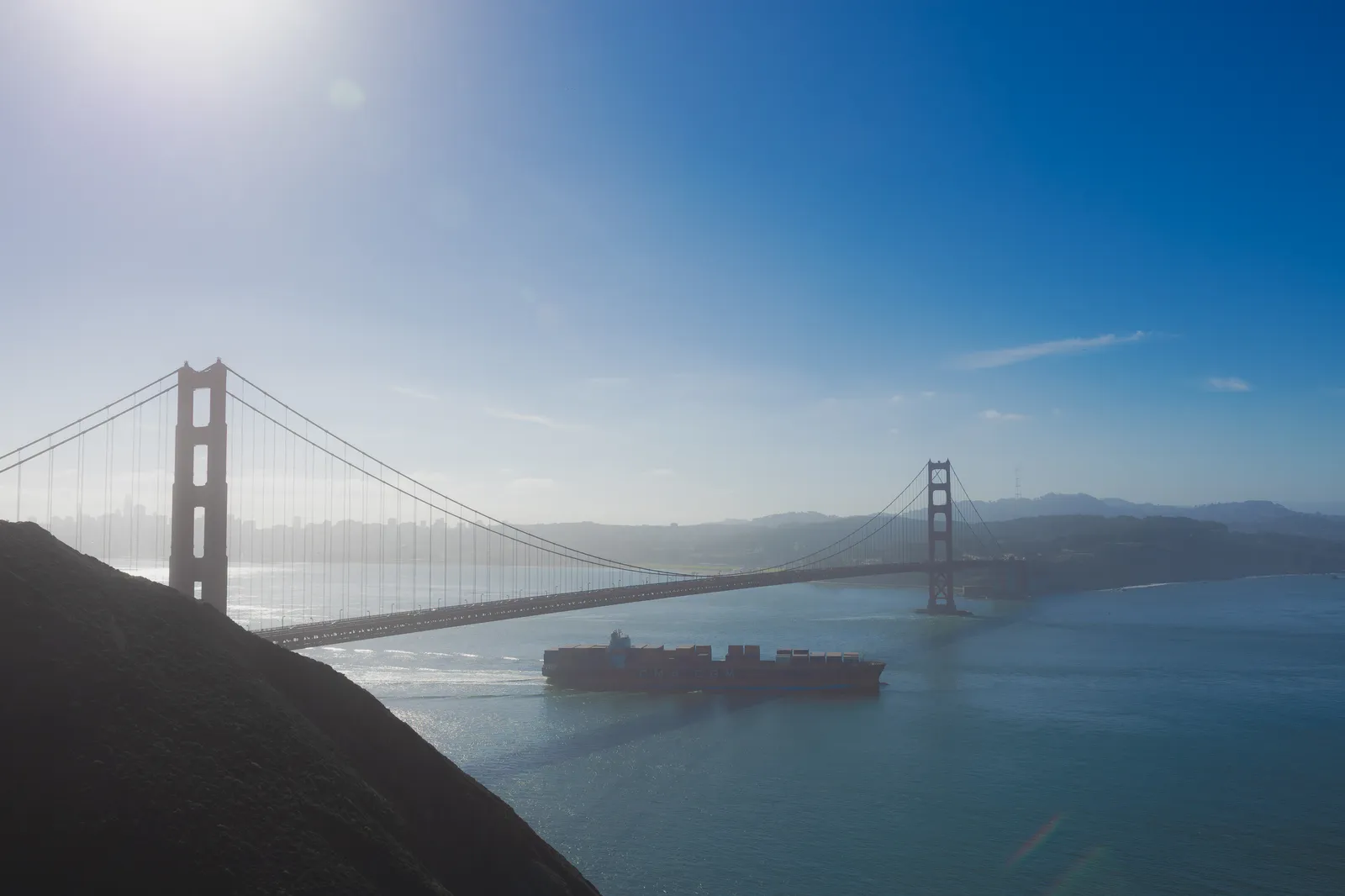 The Golden Gate Bridge stretching across the bay in hazy sunlight, with a large container ship passing beneath it and a dark hillside in the foreground.