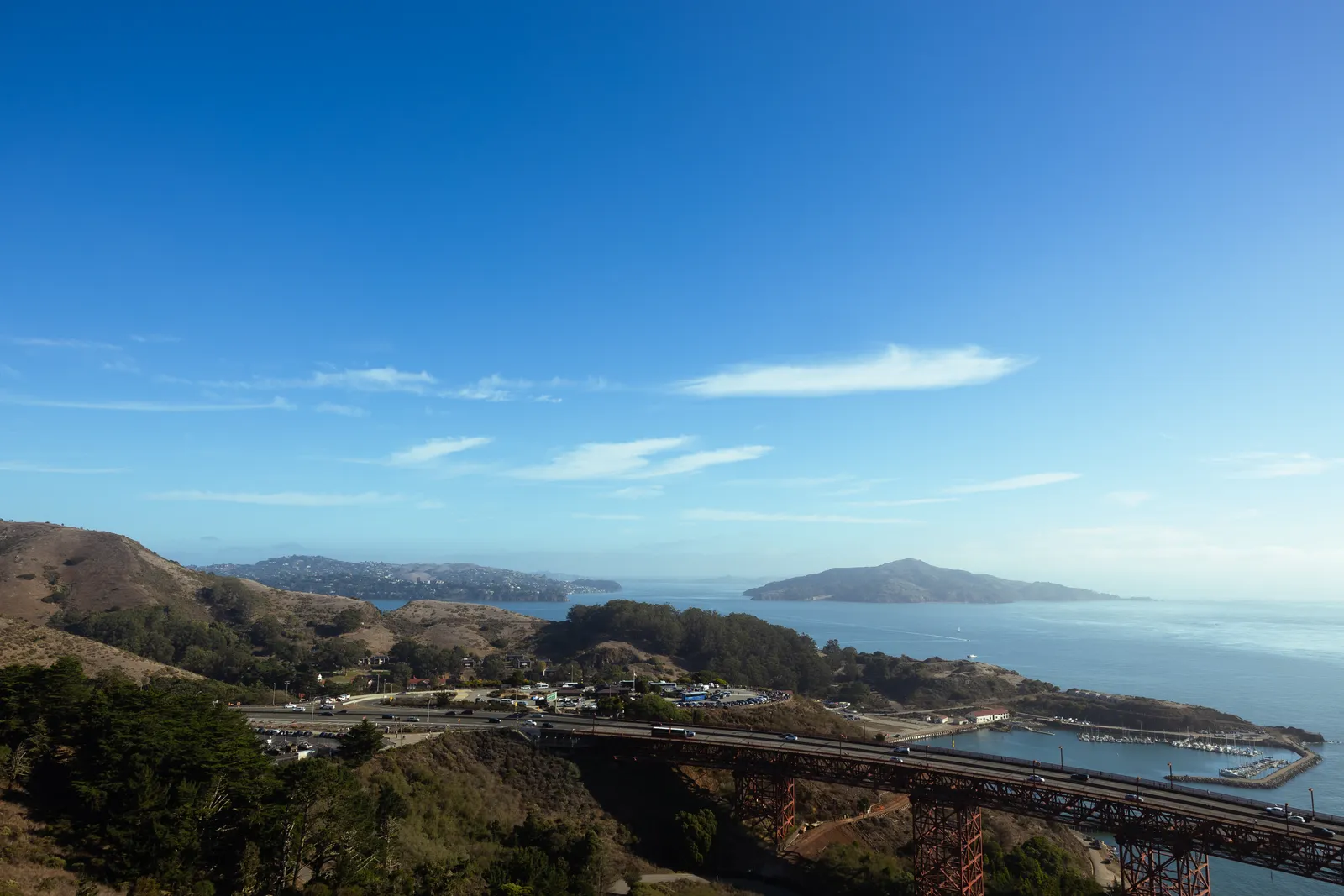 A high view over the end of the bridge, a road and a small marina beside the water, with hills, an island in the distance and a deep blue sky.