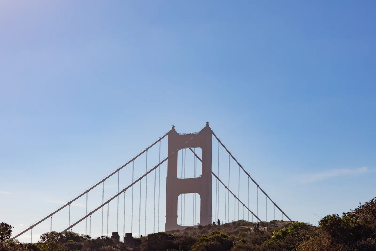 The bridge tower and cables rising above a hilltop against a clear blue sky, with a few tiny figures on the ridge below.